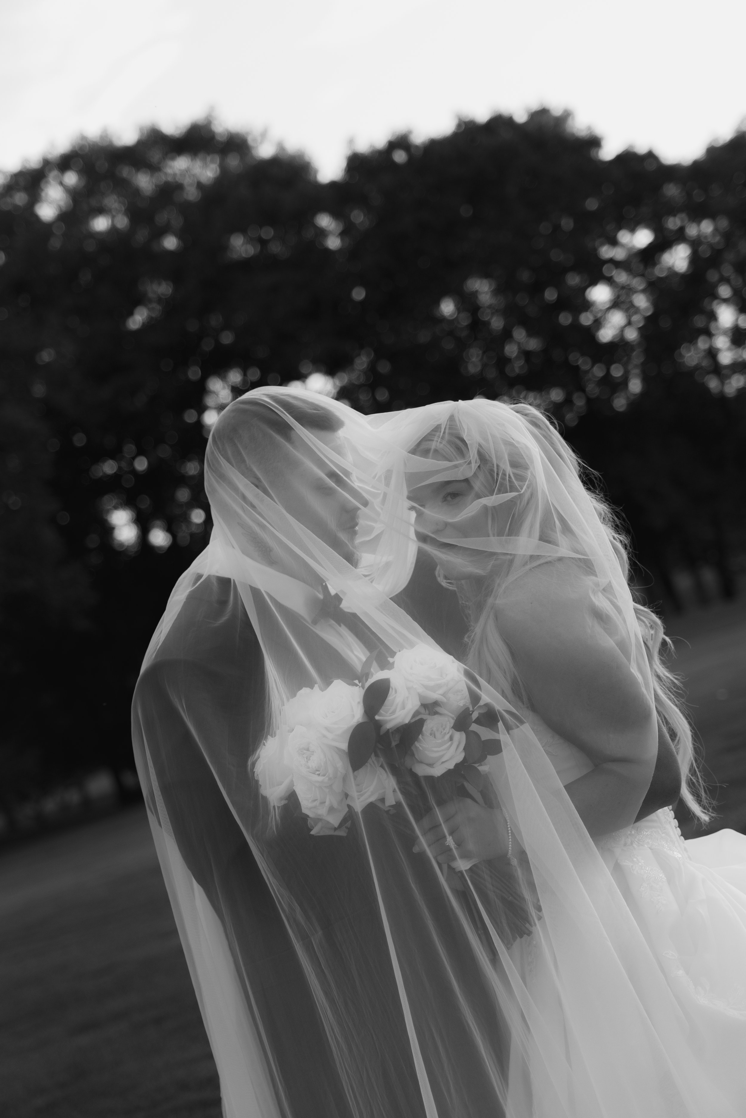 Black and white photo of two brides under a veil, holding a bouquet of white roses, with trees in the background.