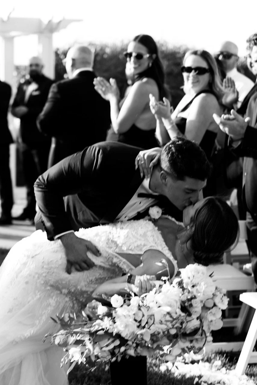 A groom leaning over and kissing the bride who is holding a bouquet of flowers during a wedding ceremony, with guests clapping and smiling in the background.