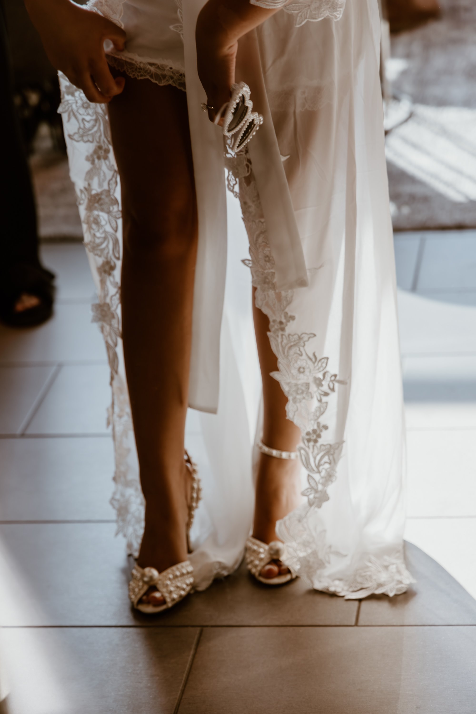 A bride adjusting her wedding dress, showing her legs and pearl-embellished high heels with lace details.