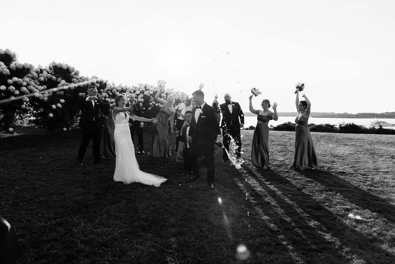 Black and white photo of a wedding celebration outdoors by a lake, with the bride and groom in the center, surrounded by friends and family, some throwing flower petals or sparklers.