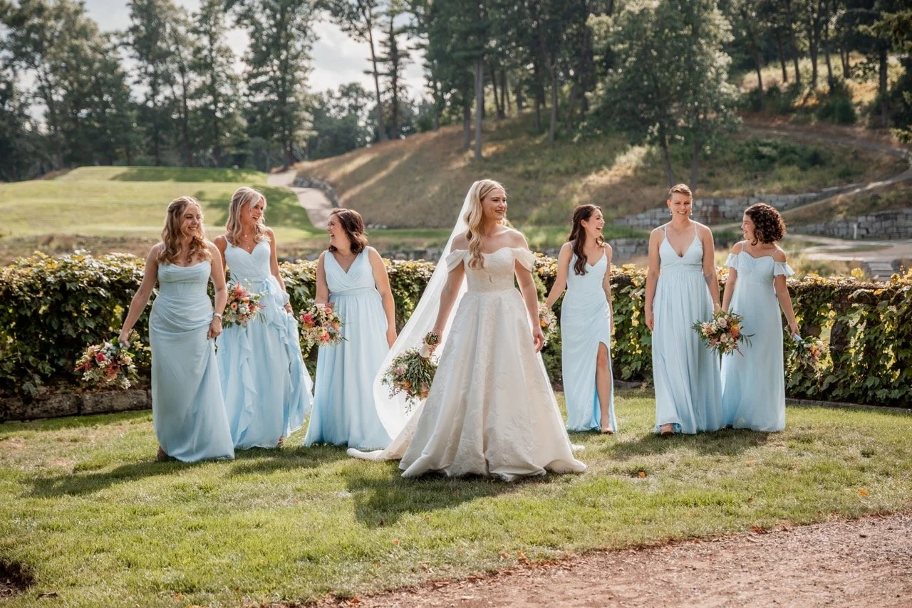 A bride in a white wedding dress and six bridesmaids in light blue dresses walking outdoors on a grassy area with trees and hills in the background.