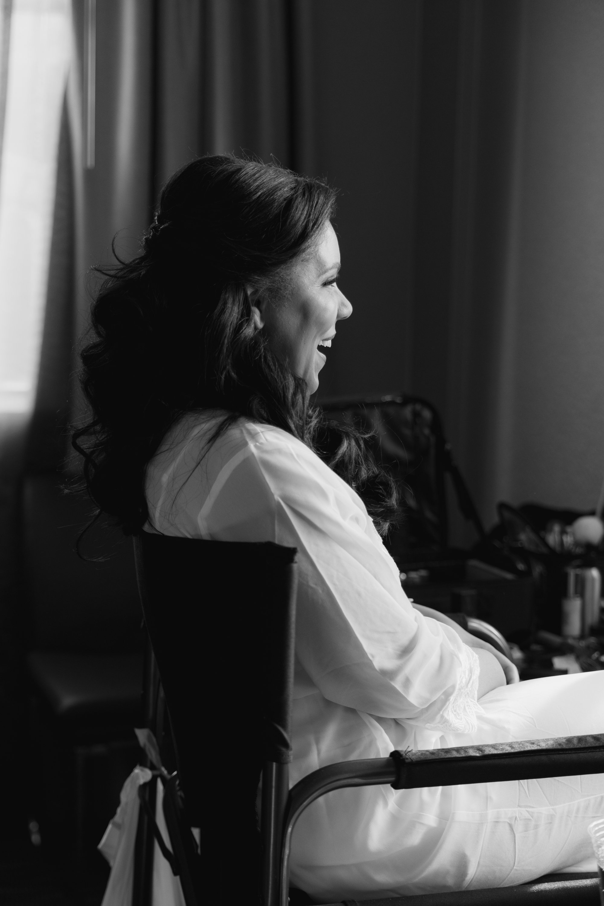A woman with long dark hair smiling and sitting in a chair, facing to the right. She is dressed in light-colored clothing, possibly getting ready for an event, with a makeup kit and other items on a table behind her.