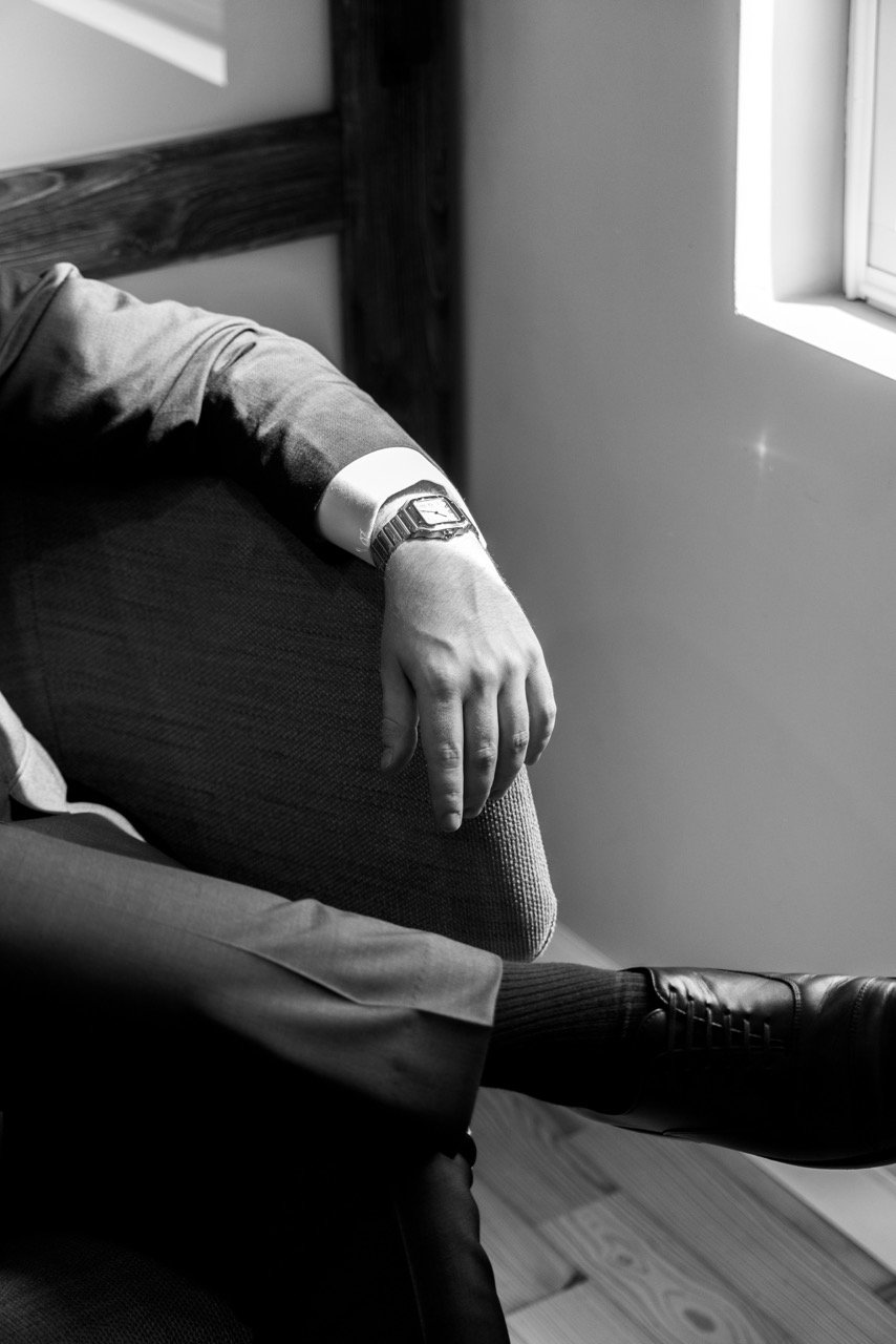 A person sitting near a window with their arm resting on a chair, wearing a wristwatch, dress shoe, and dress pants, in a black and white photo.