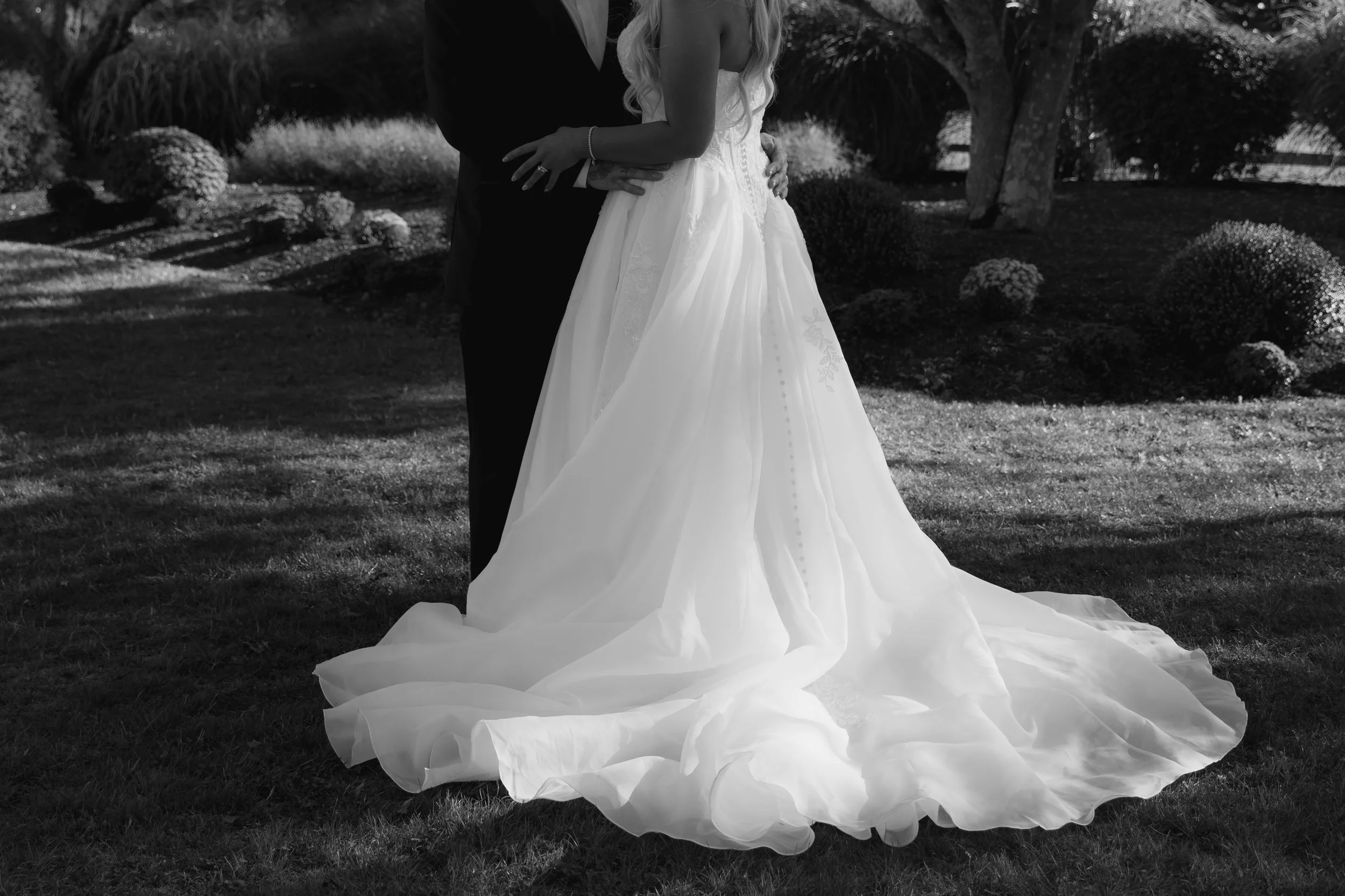 A bride and groom clothed in wedding attire holding each other outdoors, with the bride wearing a long white wedding gown and the groom in a dark suit, surrounded by trees and bushes.