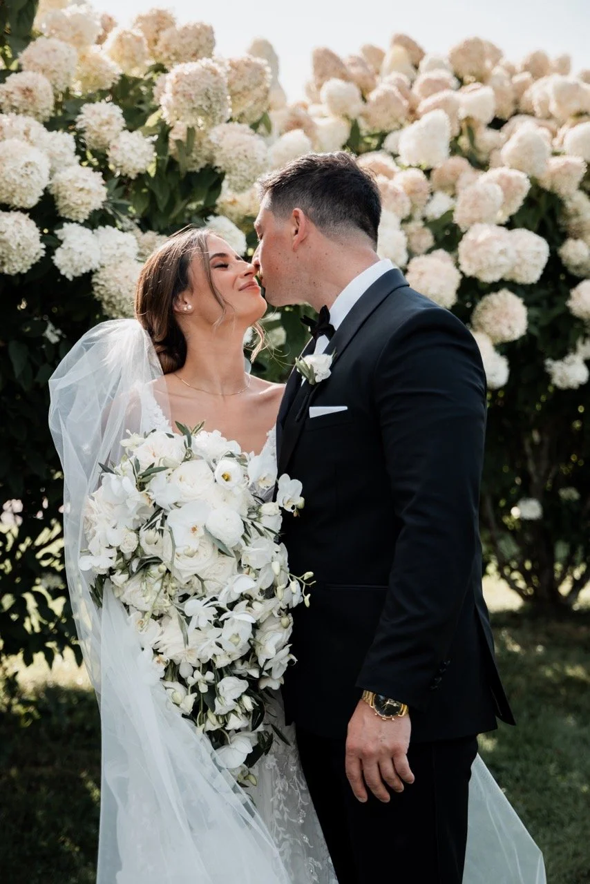 A bride and groom in wedding attire sharing a kiss outdoors. The bride holds a large bouquet of white flowers and wears a veil. They are standing in front of a bush with white flowers.