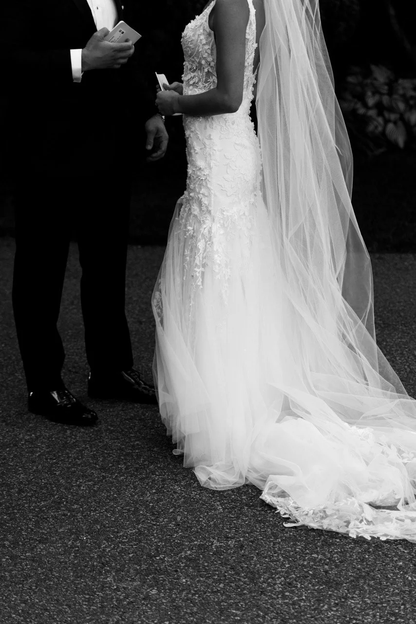 A wedding scene with a bride in a lace wedding gown and veil and a groom in a suit exchanging vows or rings, holding papers, on a dark outdoor setting.