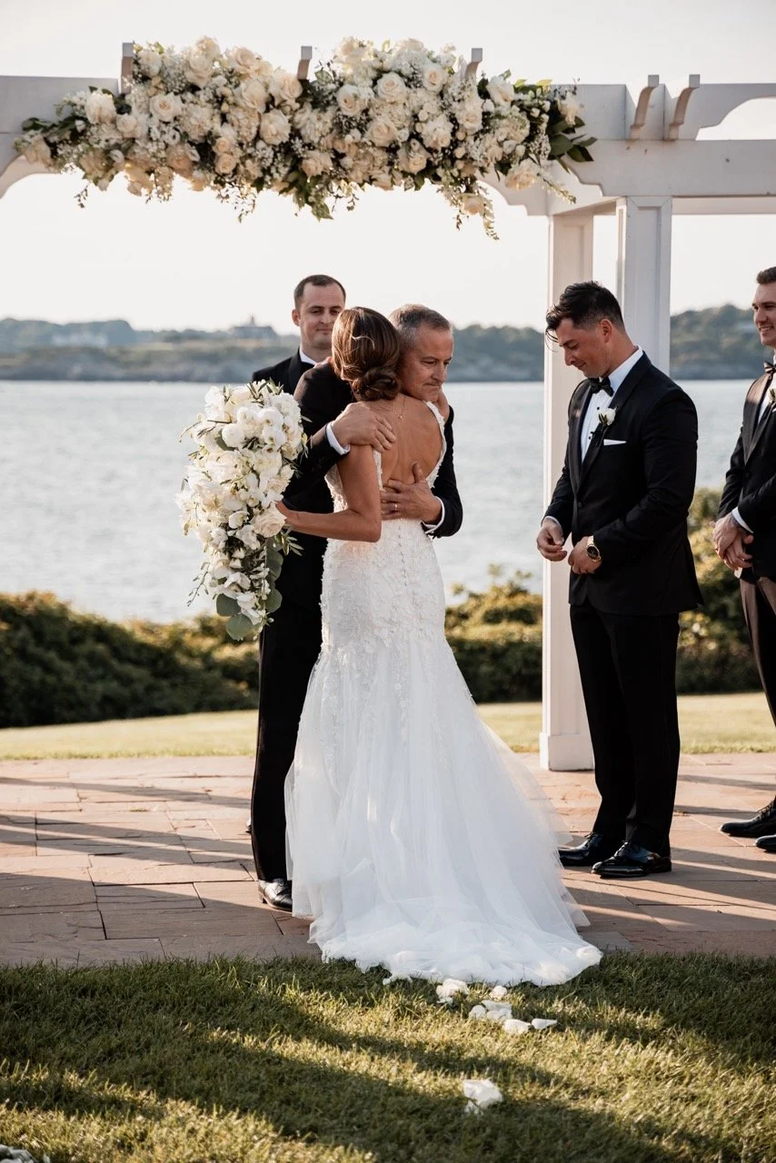A wedding ceremony taking place outdoors near a body of water, with a bride in a white gown embracing an older woman, possibly her mother, under a floral arch. The groom and groomsmen in black tuxedos stand nearby.
