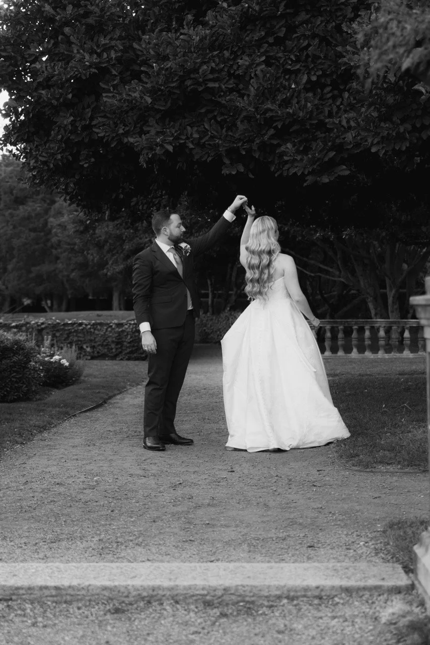 A black and white photograph of a bride and groom dancing outdoors, with the groom twirling the bride, who is wearing a flowing white wedding gown, beneath a large tree.