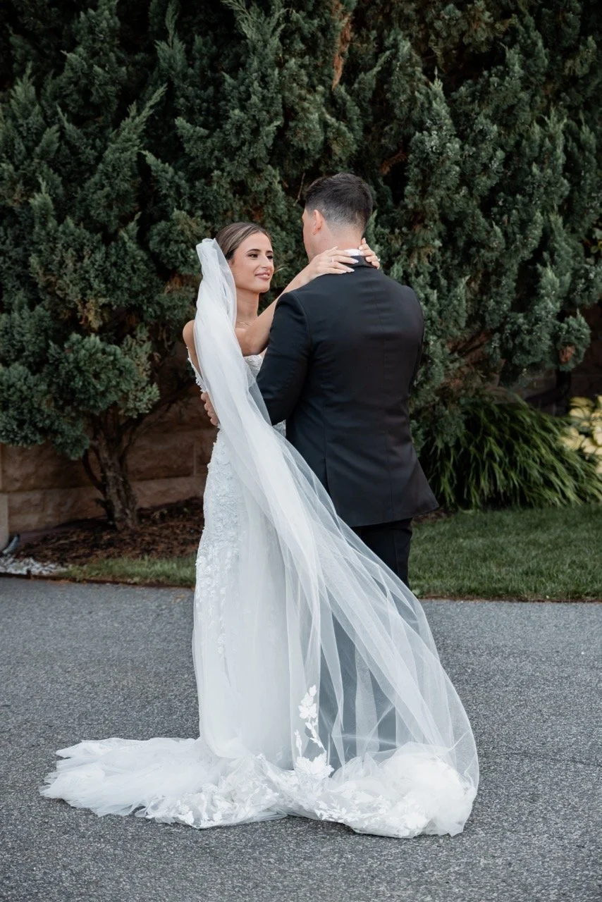 A bride and groom are dancing outdoors, with the bride wearing a white wedding gown and veil, and the groom in a black suit, surrounded by green bushes and trees.