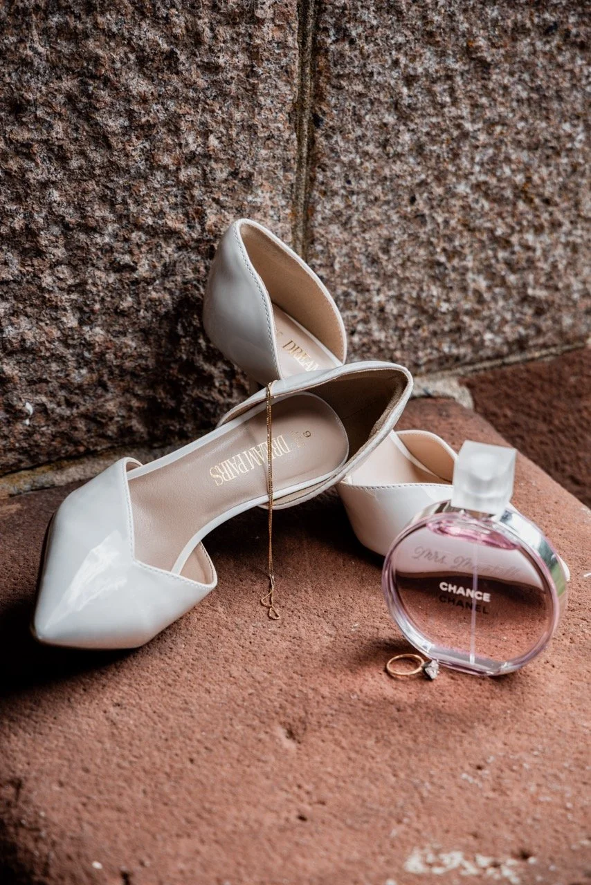A pair of white high-heeled shoes, a bottle of Chance Chanel perfume, and a delicate necklace on a brick surface.