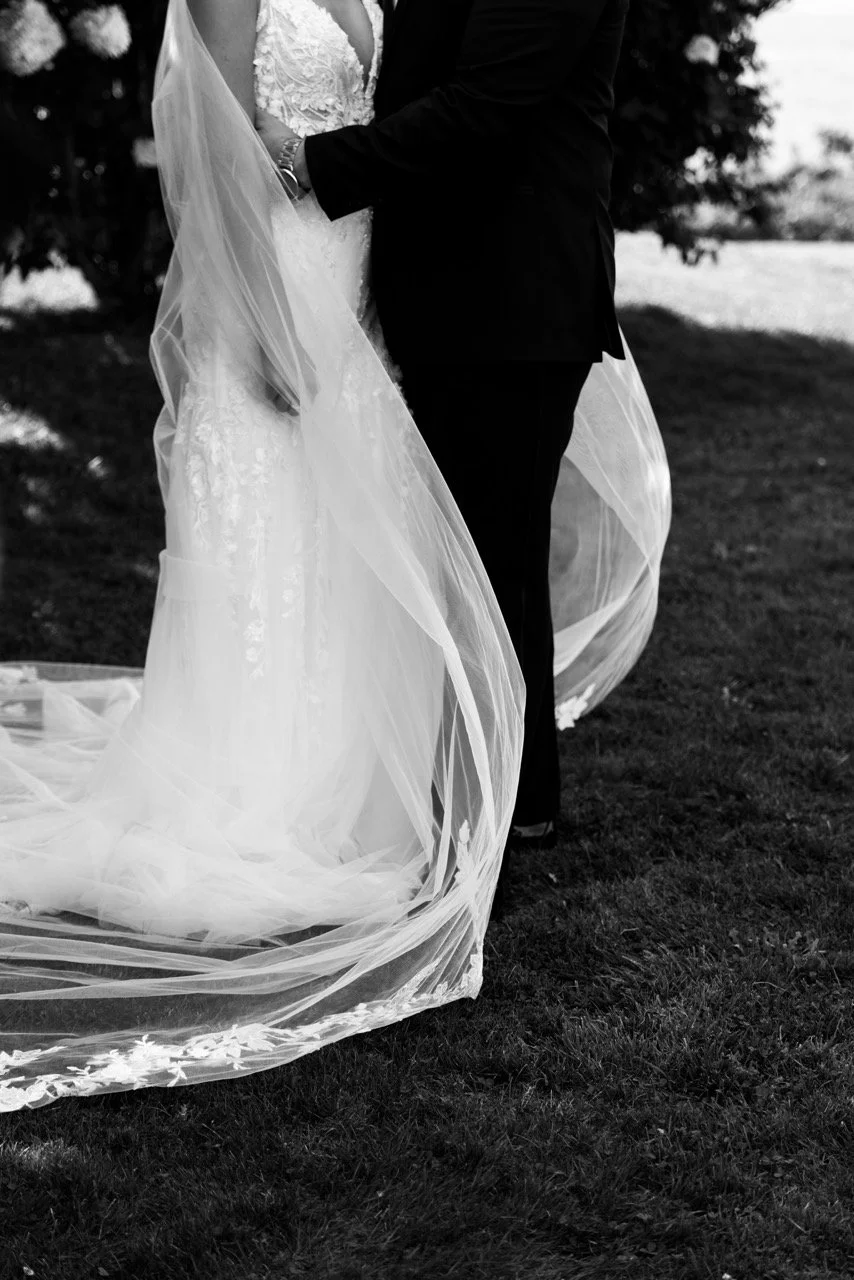 A black and white photo of a bride and groom holding hands, standing on grass outdoors, with the bride wearing a lace wedding dress and veil, and the groom in a dark suit.