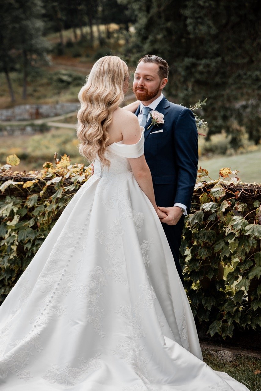 A bride and groom holding hands and looking at each other outdoors, with the bride in a white wedding gown and the groom in a navy suit, surrounded by greenery.