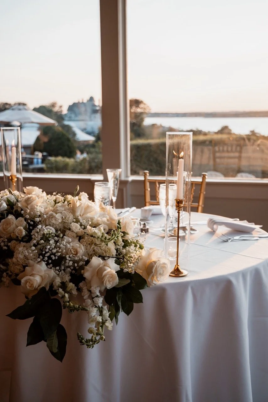 Decorated dining table with white flowers, tall candle holders with candles, and a view of a lake or river through large windows, suggestive of a wedding or special event setup.