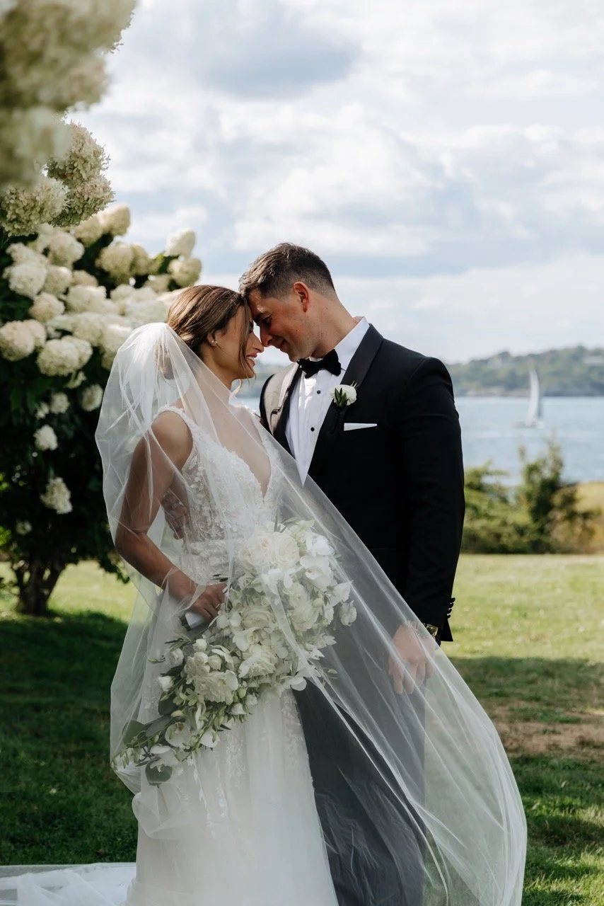 A bride and groom share a close moment outdoors, with their foreheads touching, in front of lush greenery and a body of water with sailboats in the background.