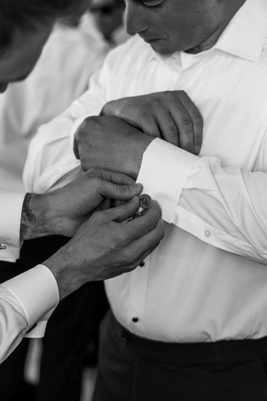 A man in a white shirt has a cufflink being fastened by another person in a white shirt, in a black and white photo.