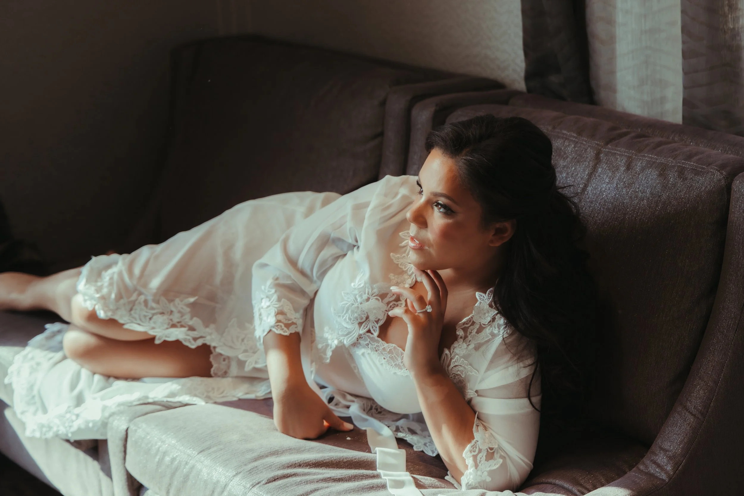 A woman in a white lace robe lying on a dark sofa, looking thoughtfully to the side with her hand near her chin.