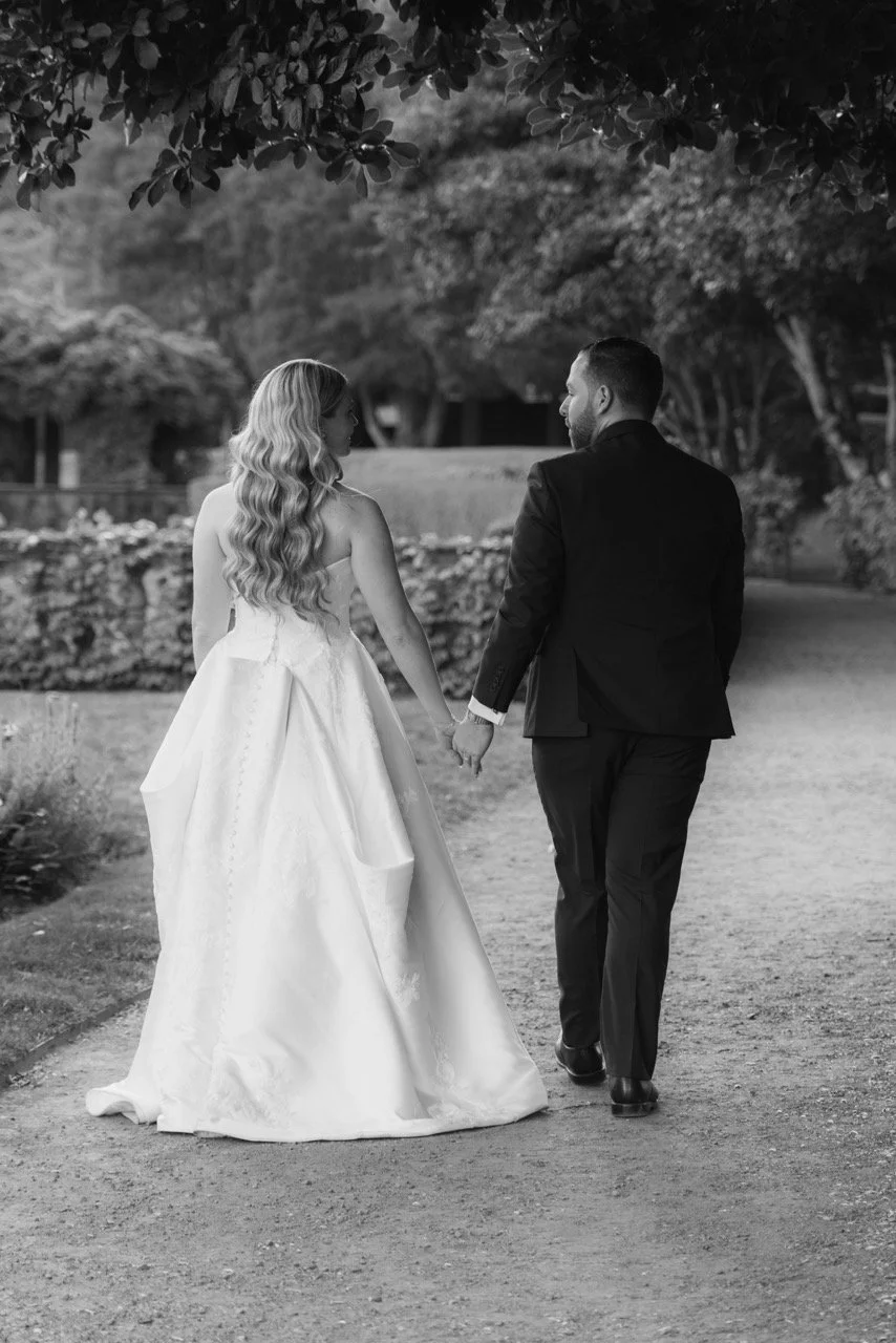 Black and white photo of a bride and groom holding hands and walking away in a park or garden. The bride has long wavy hair and is wearing a sleeveless wedding gown. The groom has short hair and is wearing a suit. They are looking at each other as th