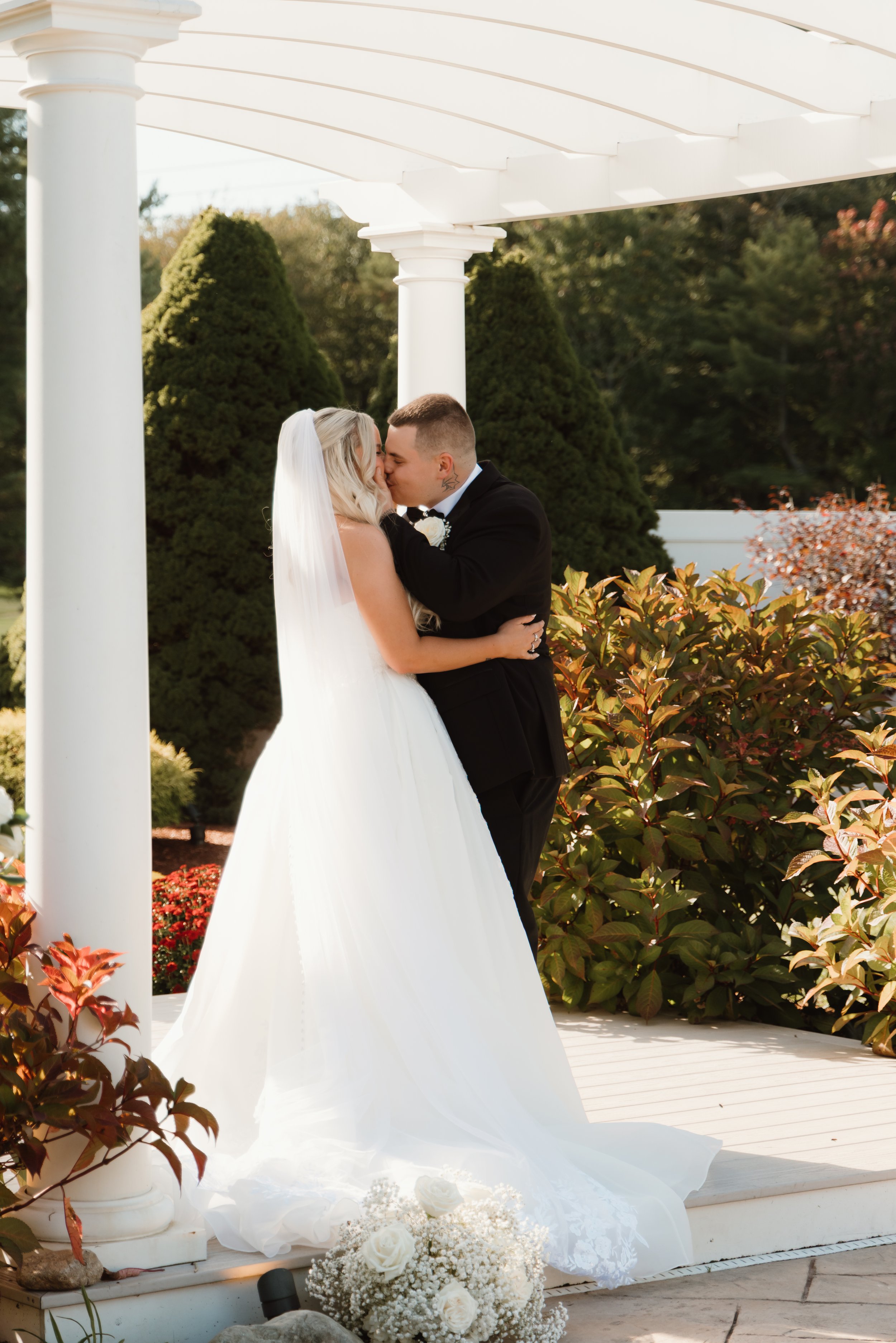 A bride and groom kiss under a white pergola on a sunny outdoor wedding day, surrounded by green shrubbery and plants.