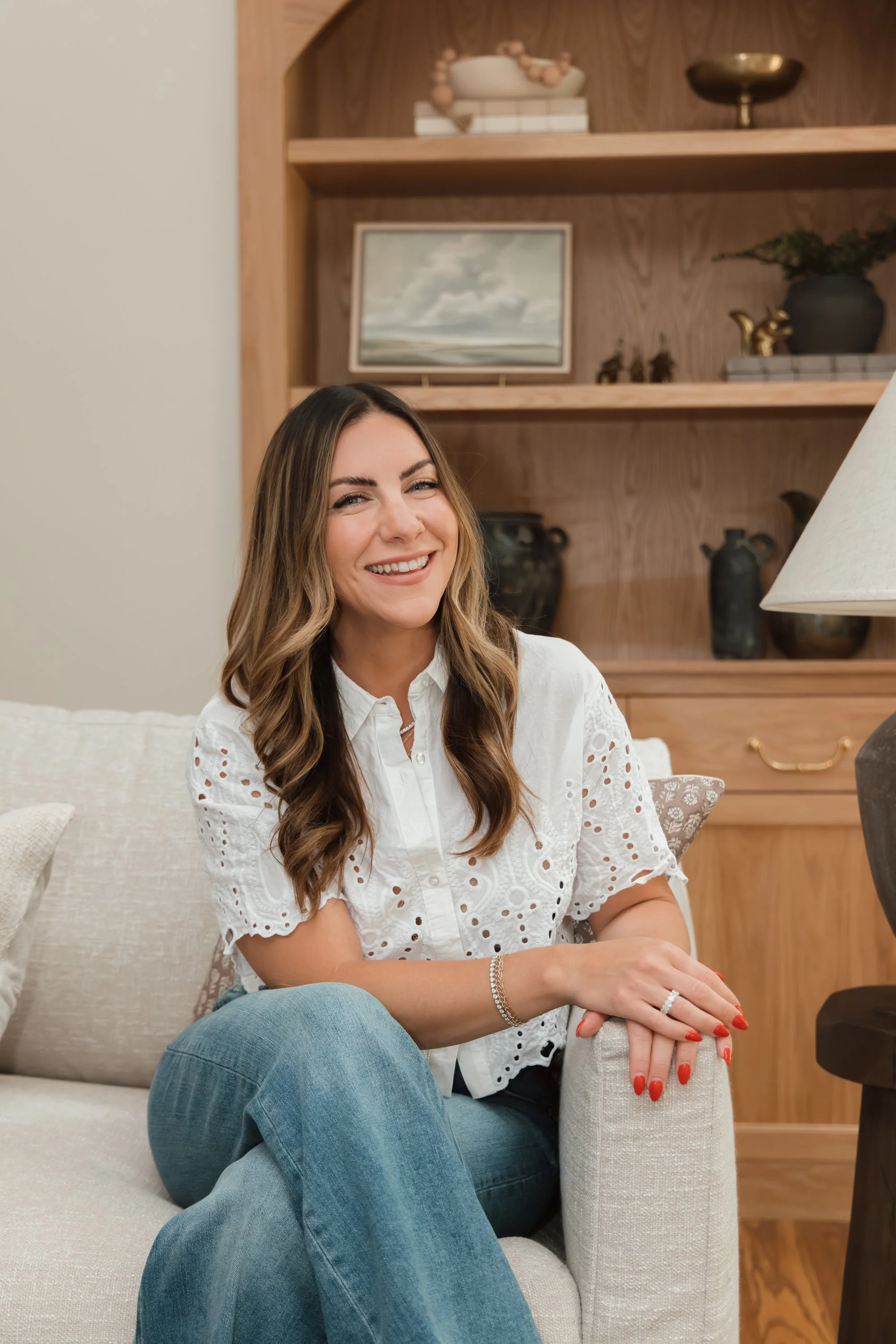 A woman with long brown hair, smiling, wearing a white eyelet blouse and blue jeans, sitting on a beige sofa in a living room.