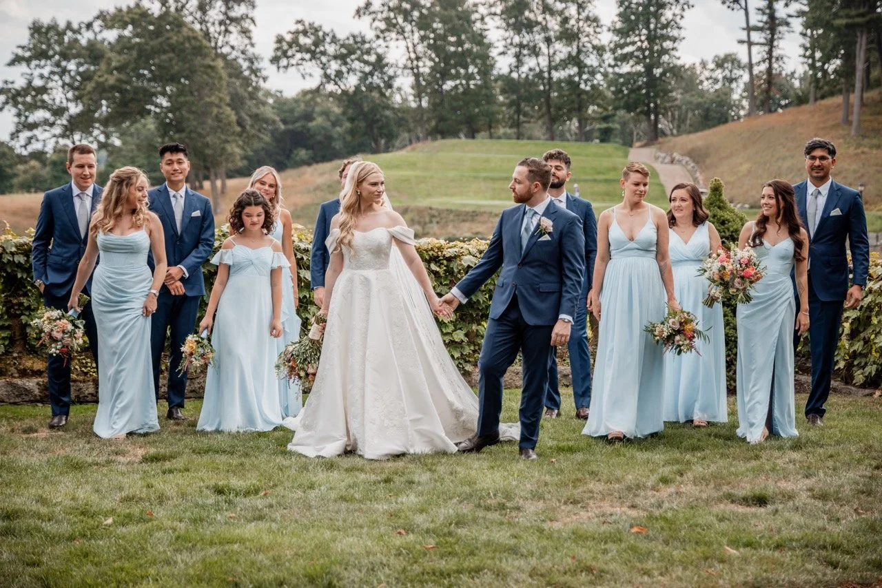 A wedding party outdoors, with the bride and groom holding hands in center, surrounded by bridesmaids and groomsmen, all dressed in elegant attire, with greenery and trees in the background.