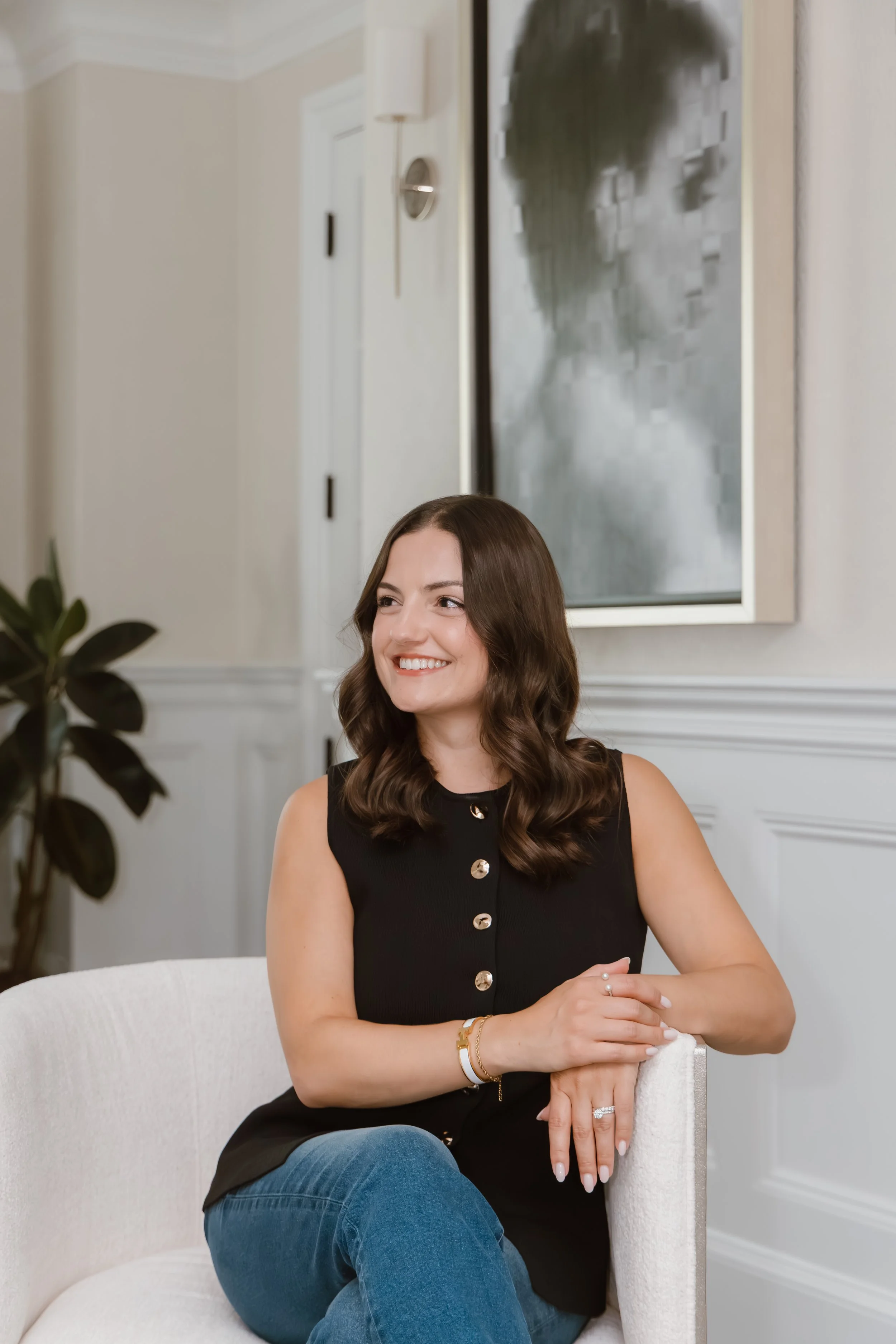 A woman with long brown wavy hair smiling while sitting on a white chair in a room with white paneled walls and a large metallic abstract wall art