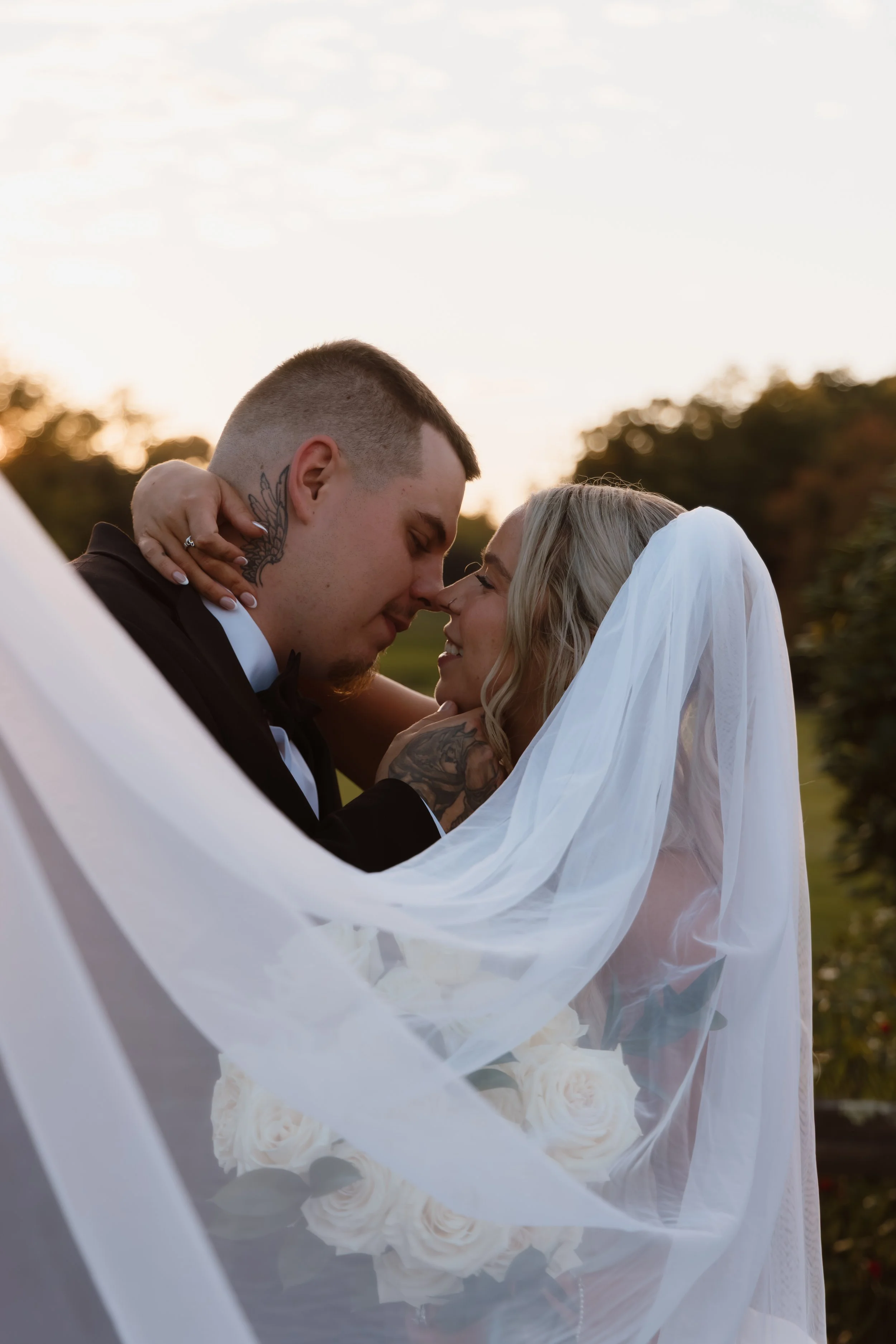 Bride and groom sharing a romantic moment, with their foreheads touching, at sunset outdoors, wrapped in white wedding veil and bouquet of white roses.