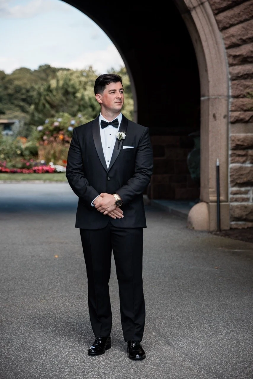 A man in a black tuxedo with a bow tie, white dress shirt, and boutonniere stands with his hands clasped in front outdoors near an arched stone structure, with trees and flowers in the background.