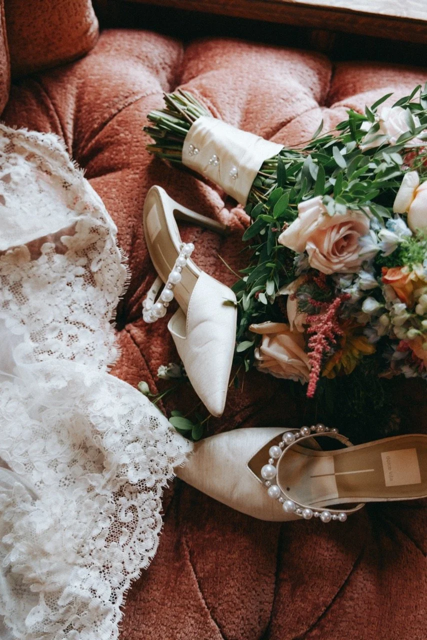 A pair of white high-heeled shoes with pearl embellishments, a bouquet of pink and white flowers, and a piece of white lace fabric arranged on a vintage pink armchair.