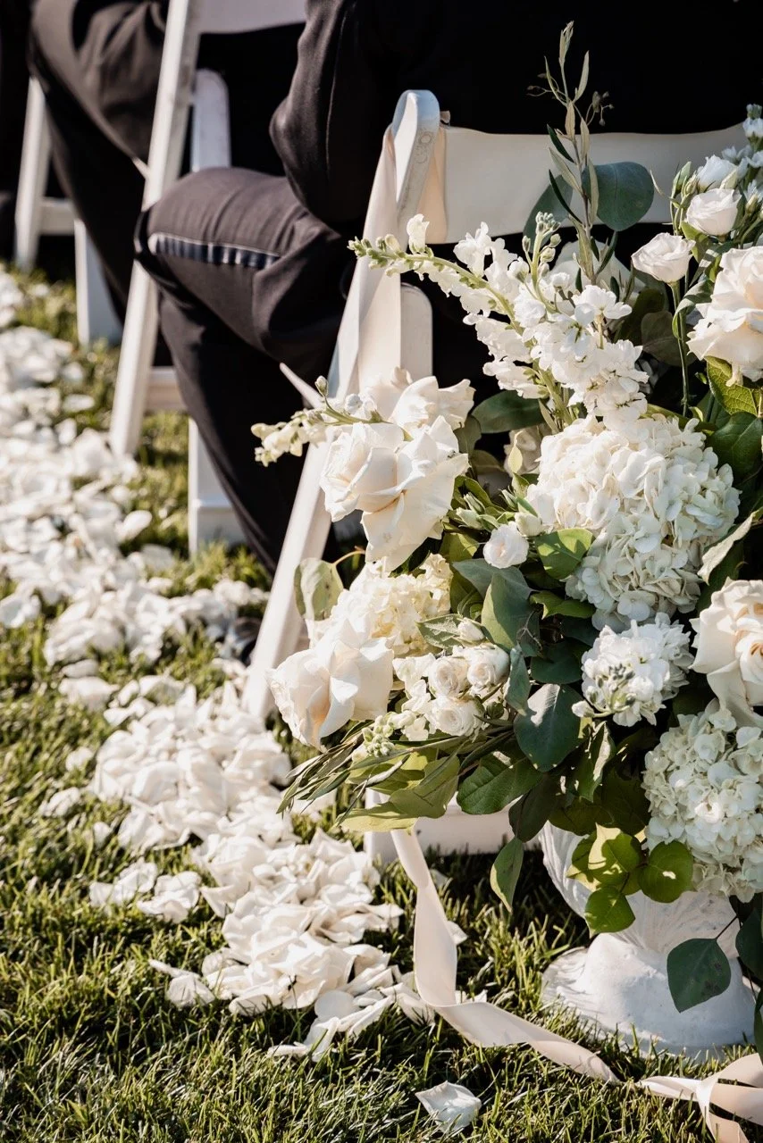 A floral arrangement of white flowers such as roses and hydrangeas with greenery, placed next to white chairs at an outdoor wedding ceremony, with flower petals scattered on the grass.