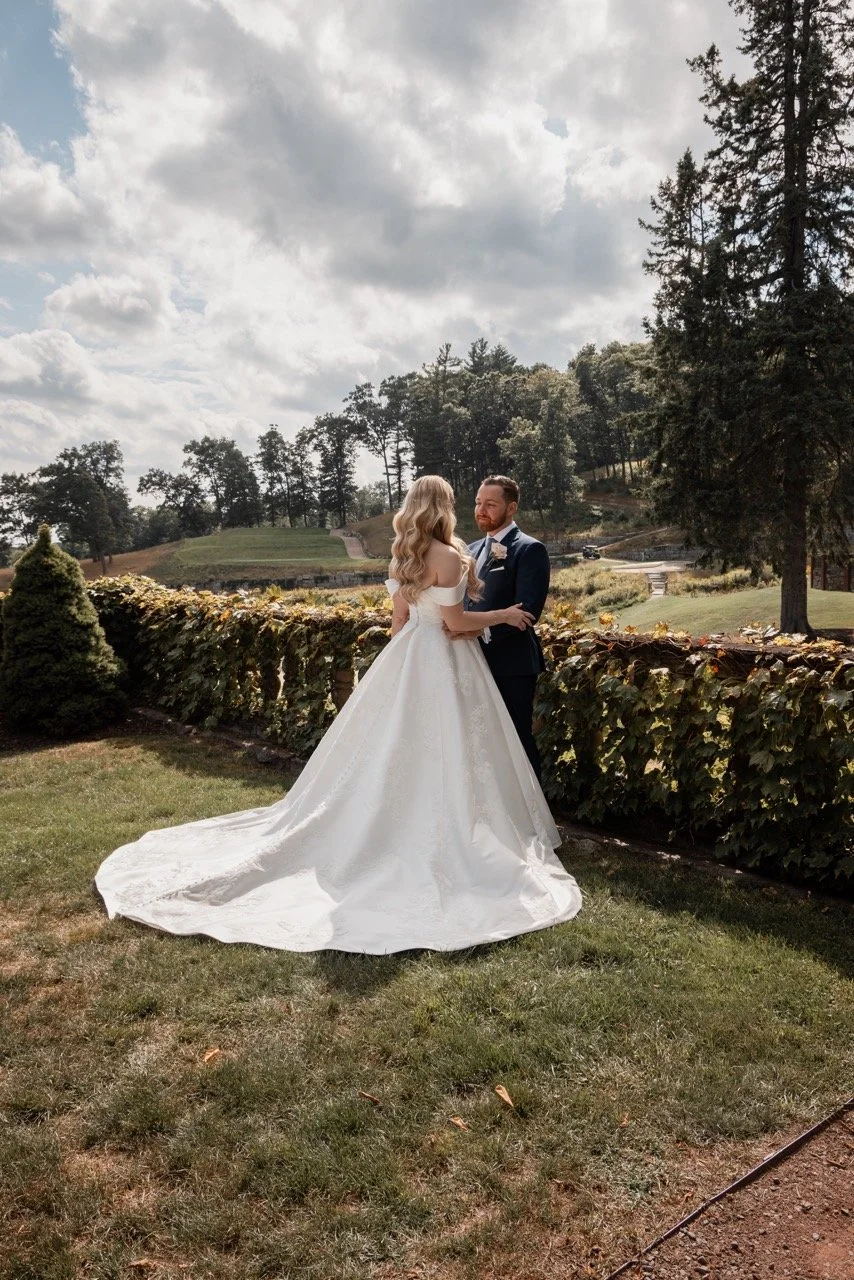 Bride and groom holding hands outdoors on a grassy area with trees and shrubs in the background under a partly cloudy sky.
