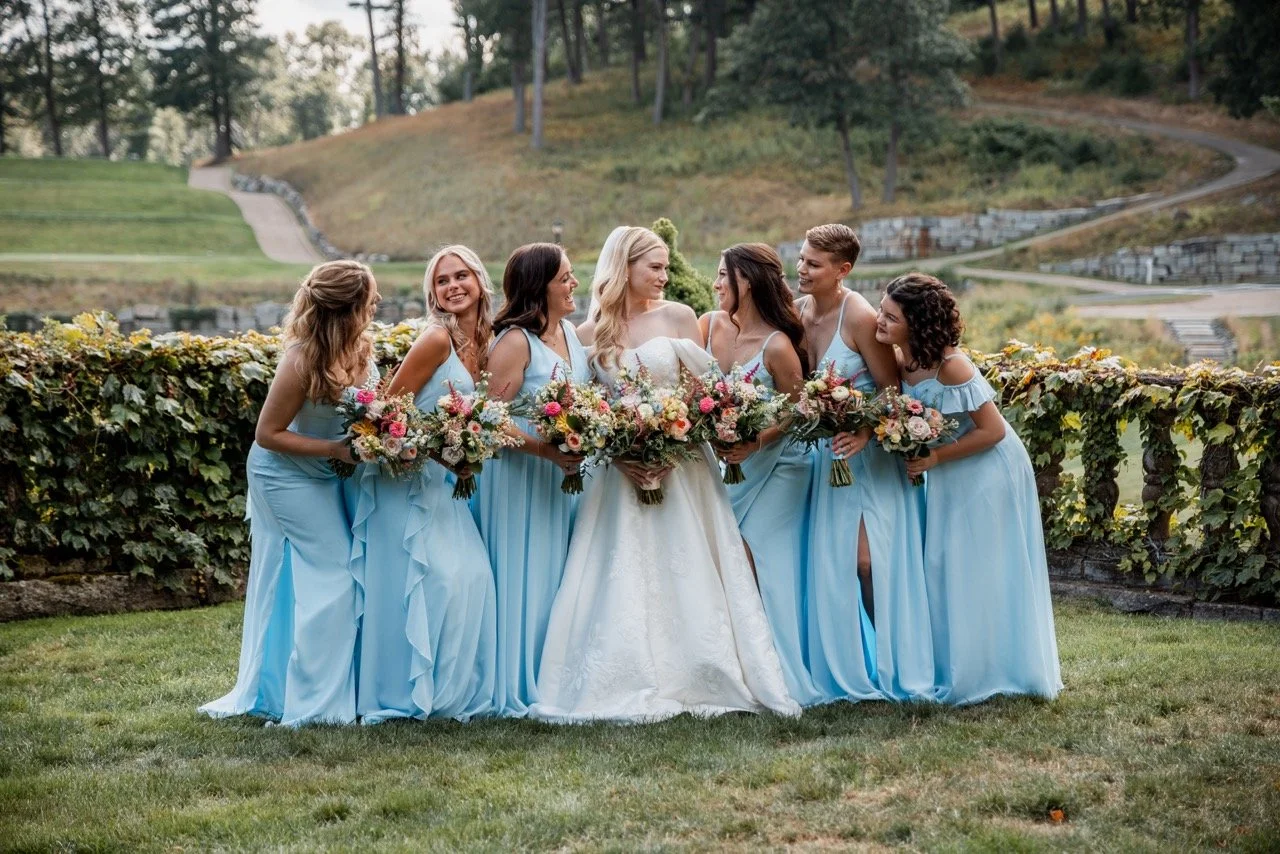 A bride and six bridesmaids standing outdoors in a garden, holding bouquets of flowers, with a scenic background of trees and hills.