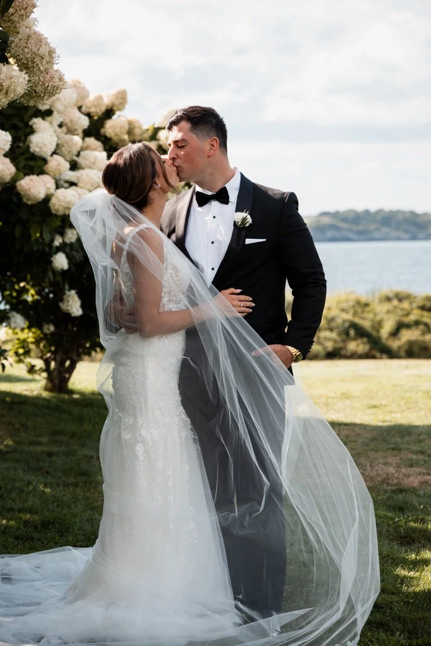 A bride and groom sharing a kiss outdoors on their wedding day, with the bride in a white gown and veil, and the groom in a black tuxedo, near a bush with white flowers and a body of water in the background.