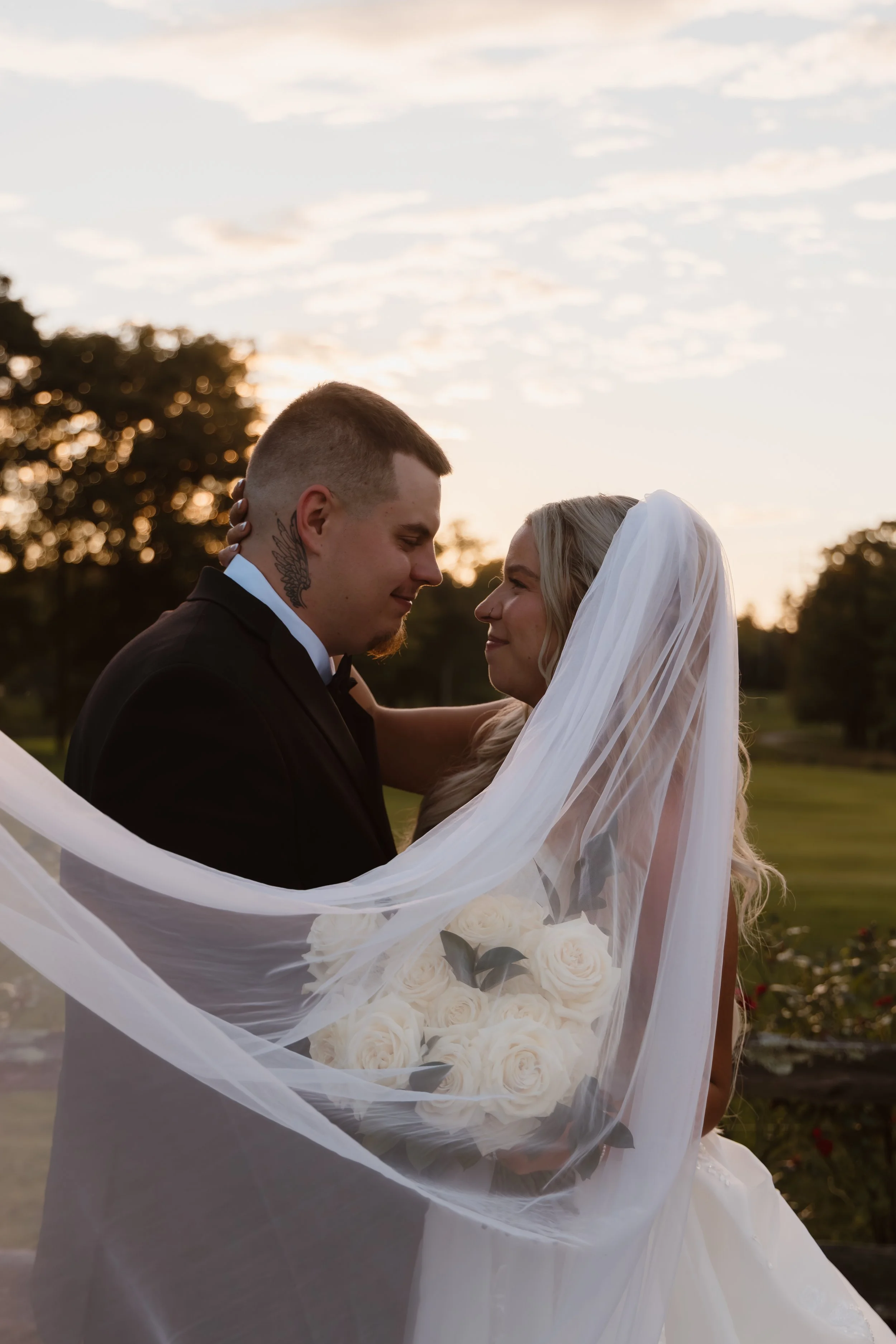 A bride and groom sharing a kiss at sunset, with the bride holding a bouquet of white roses and her veil flowing in the breeze.