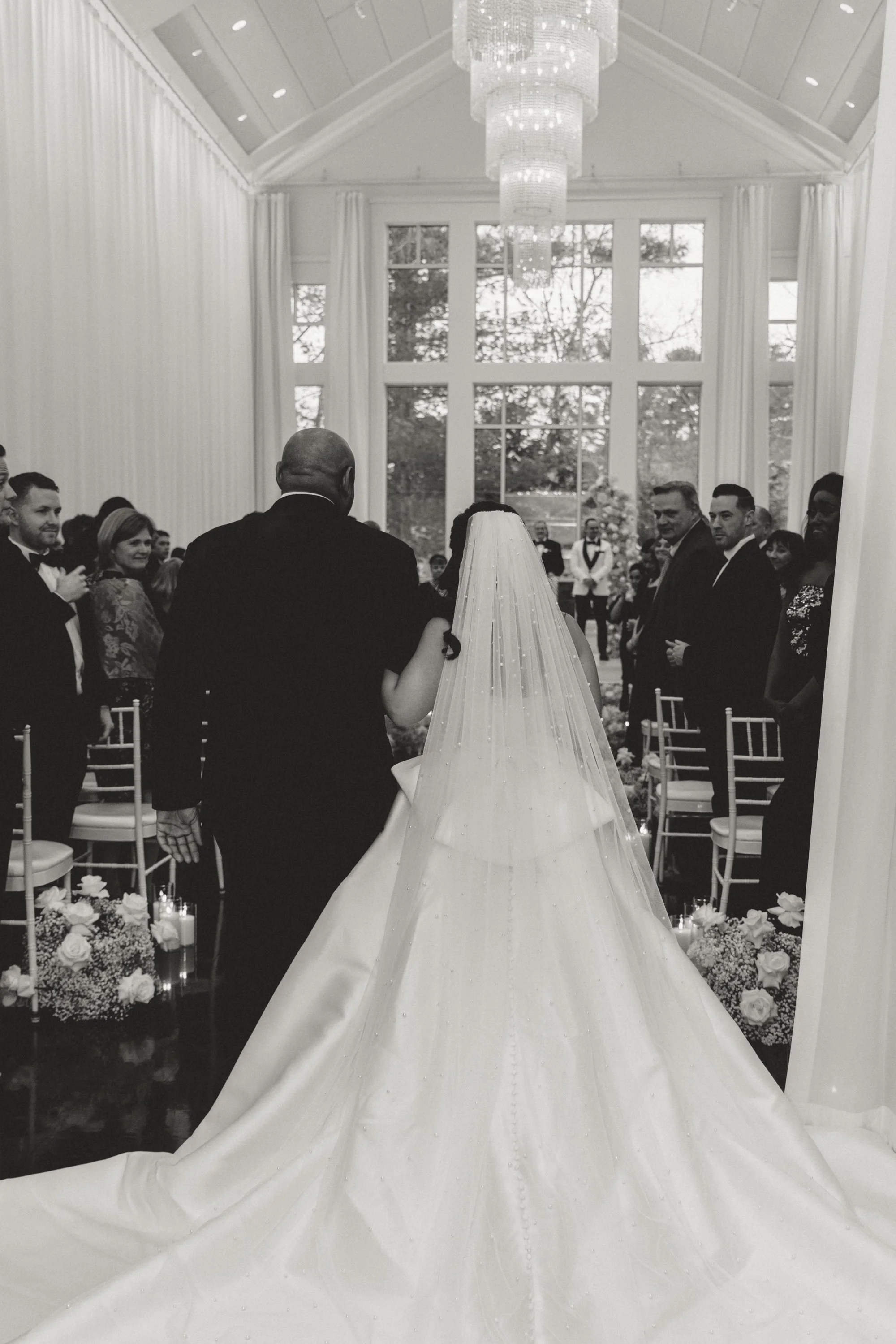 Bride in a wedding gown walking down the aisle, flanked by seated guests, in a bright indoor venue with large windows and a chandelier.