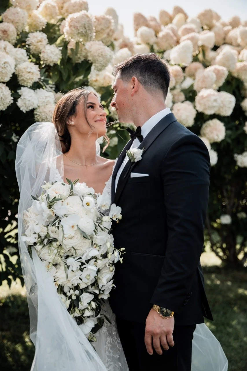 A bride and groom facing each other outdoors, surrounded by white and pale pink floral bushes, with the bride holding a large bouquet of white flowers. The bride wears a white wedding gown and veil, and the groom is in a black tuxedo with a bow tie.