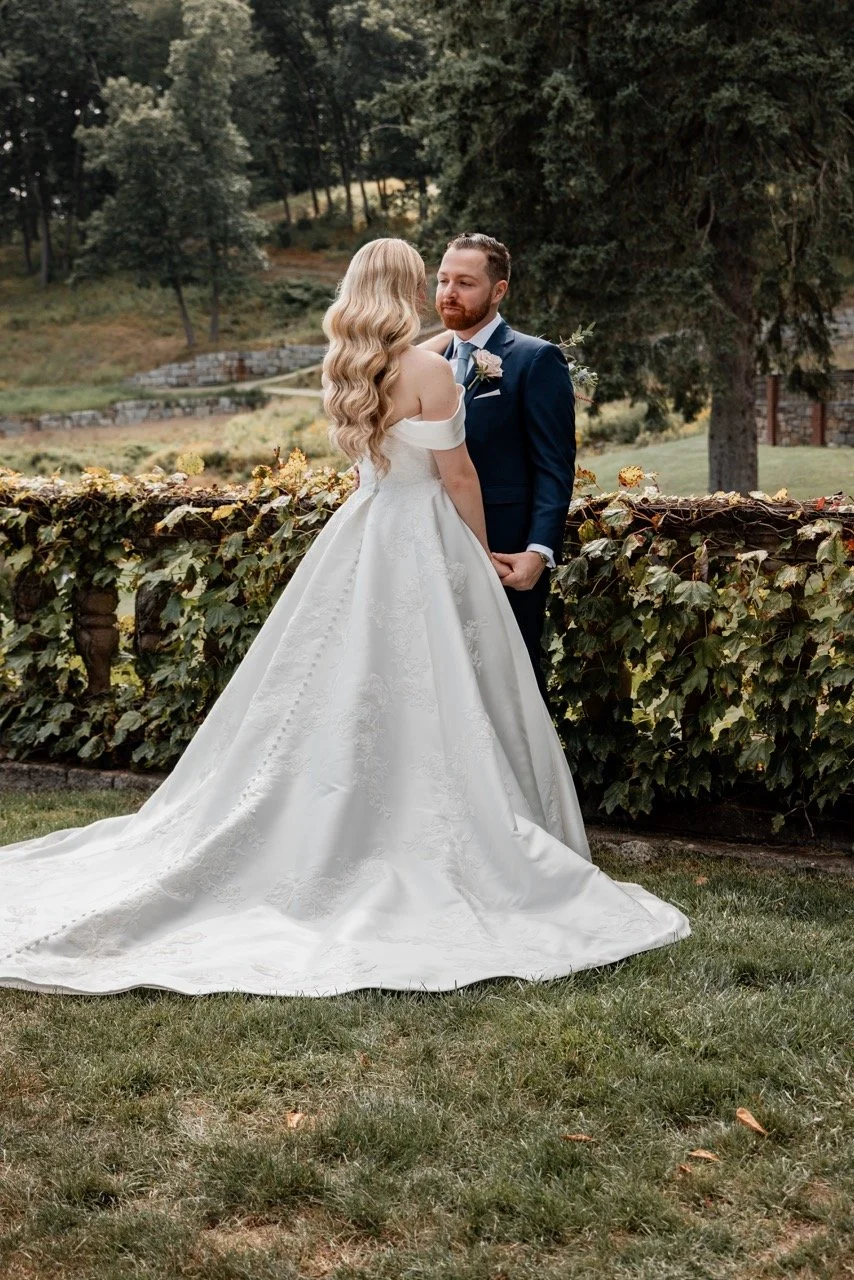 A bride and groom standing outdoors in a garden, facing each other, surrounded by greenery and trees, with the bride wearing a white off-the-shoulder wedding dress and the groom wearing a navy blue suit with a boutonniere.