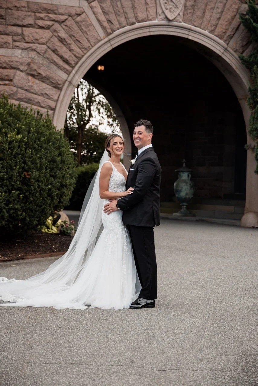 A bride and groom smiling and embracing in front of a stone archway at sunset.