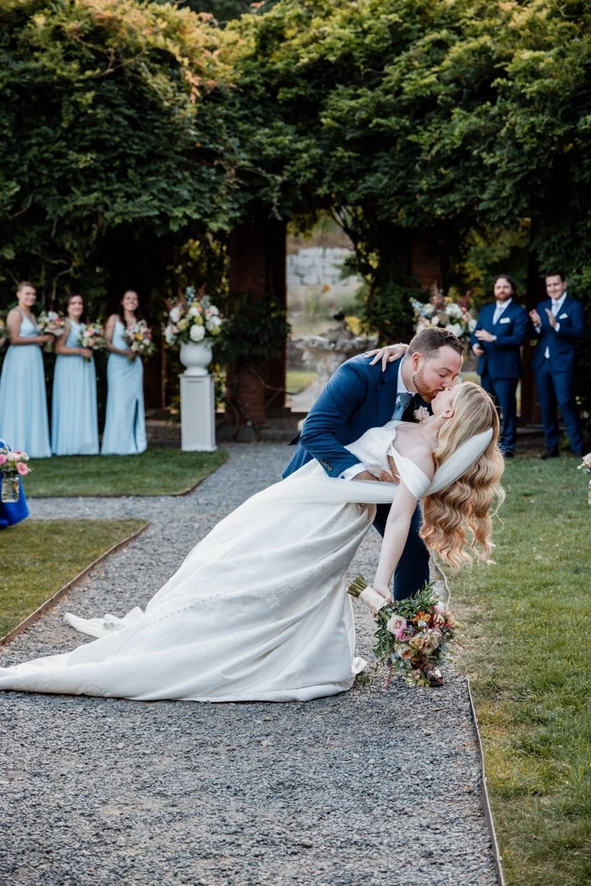 A newlywed couple shares a romantic dip kiss during their outdoor wedding ceremony, with wedding party members and lush greenery in the background.
