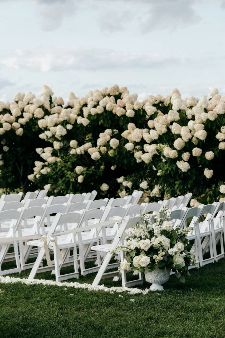 Wedding ceremony setup with white folding chairs arranged outdoors on grass, decorated with a large floral arrangement in a white vase, and white flower petals scattered on the ground. In the background, white hydrangea bushes line the scene, with a 