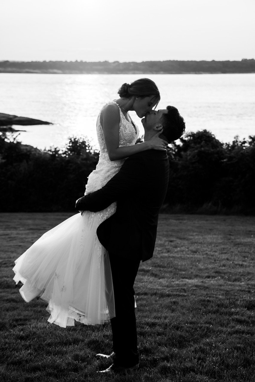 A couple in wedding attire sharing a kiss outdoors near a body of water, with trees and the water in the background, captured in black and white.