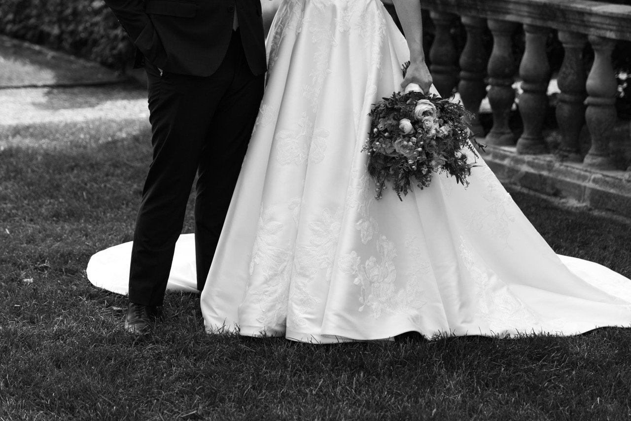 Black and white photo of a bride and groom standing outside on grass, with the bride holding a bouquet, next to a stone railing.