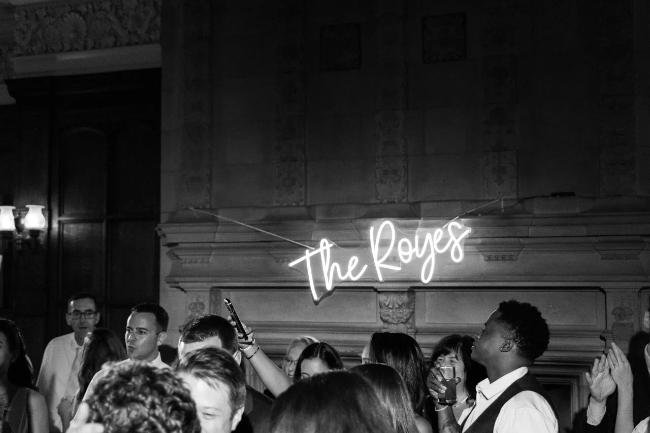 People at a social gathering with a neon sign reading 'the Royers' in a dimly lit room with wood-paneled walls.