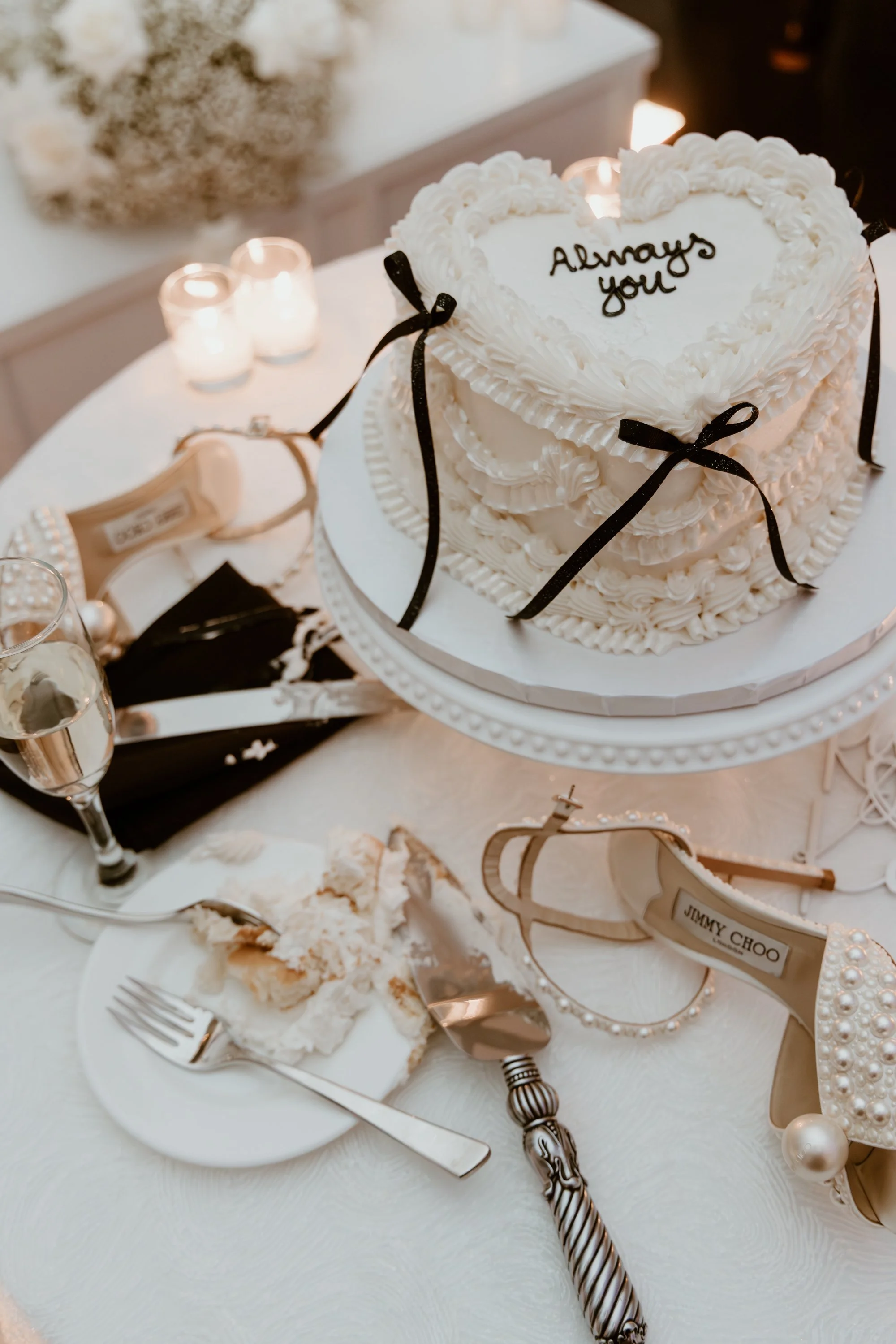 A white wedding cake with piped frosting, decorated with black ribbons, and the words 'Always you' written on top. The cake is on a white table with a champagne glass, a half-eaten cake slice, a wooden cake server, and a pair of pearl-embellished sun