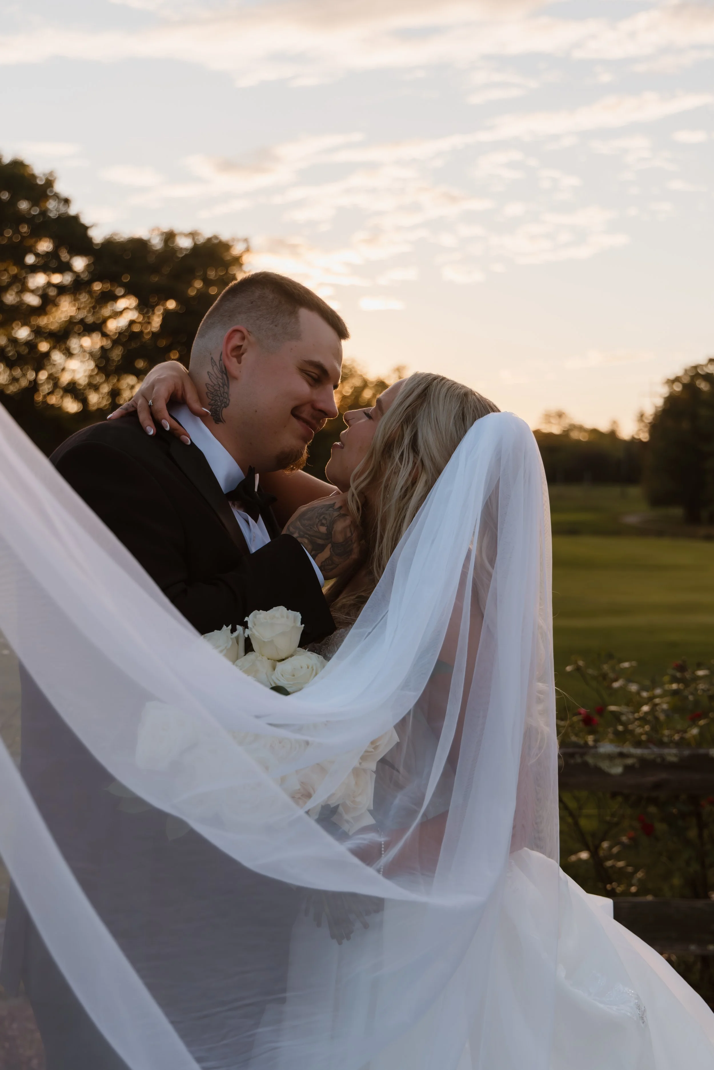 A newlywed couple sharing a loving moment outdoors at sunset, with the bride wearing a veil and holding a bouquet of white roses.