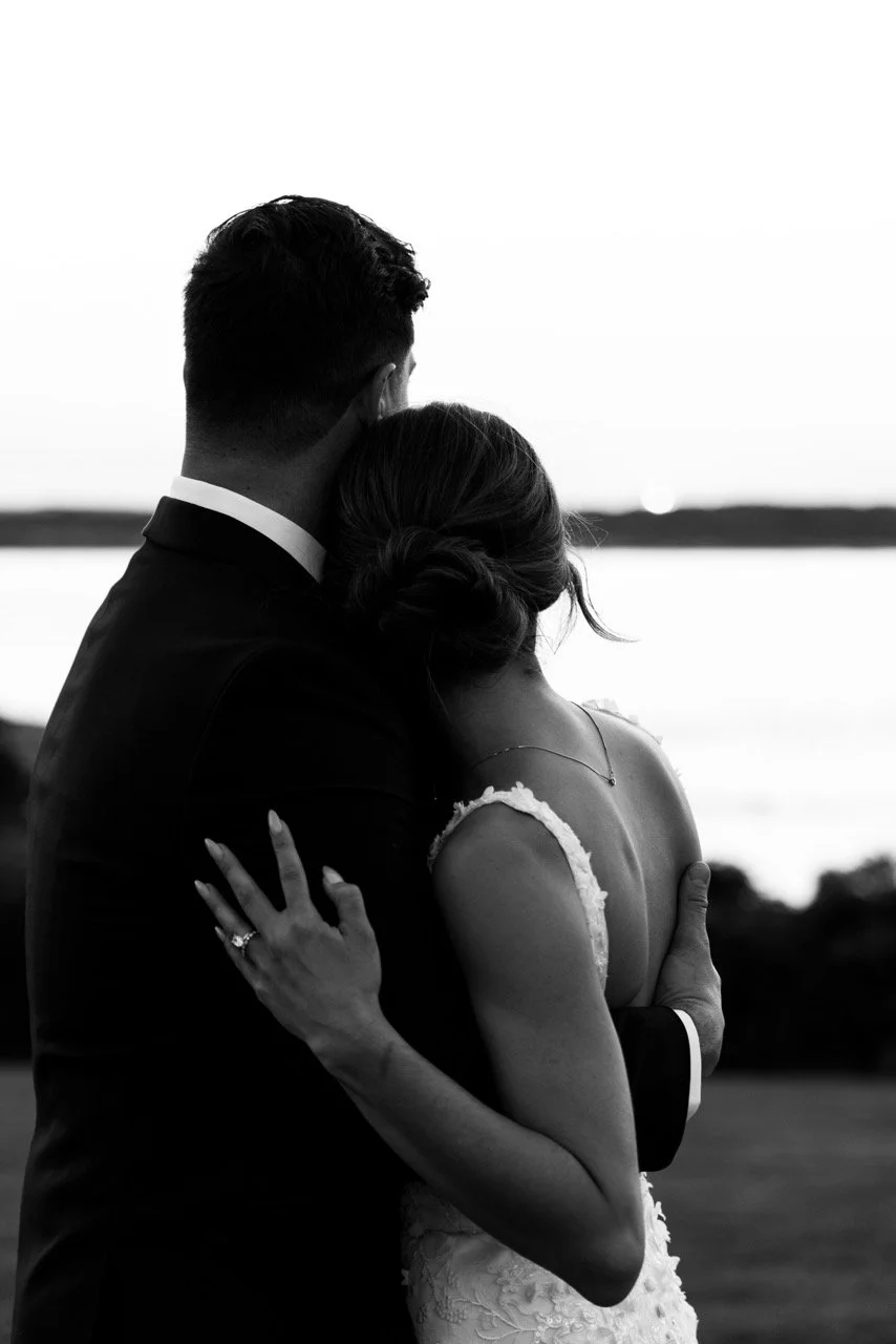 A black and white photo of a couple embracing outdoors, with a body of water and horizon in the background.