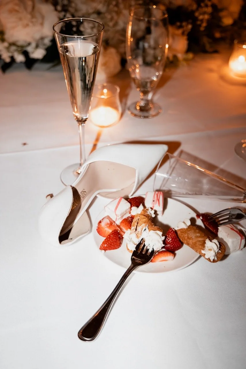A partially eaten plate of strawberry shortcake with whipped cream and strawberries, two forks, a glass lying on its side, a champagne flute, and a wine glass on a table with wine glasses and candles in the background.