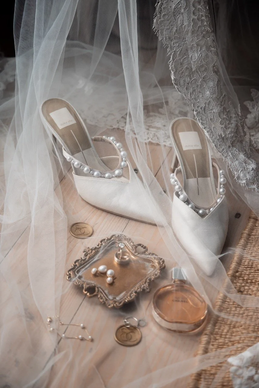 A pair of white high-heeled shoes decorated with large pearl beads inside, placed under a sheer veil or fabric. Surrounding items include a small ornate silver tray with pearl jewelry, a round pinkish jar, two gold coins, and a pair of earrings, all 