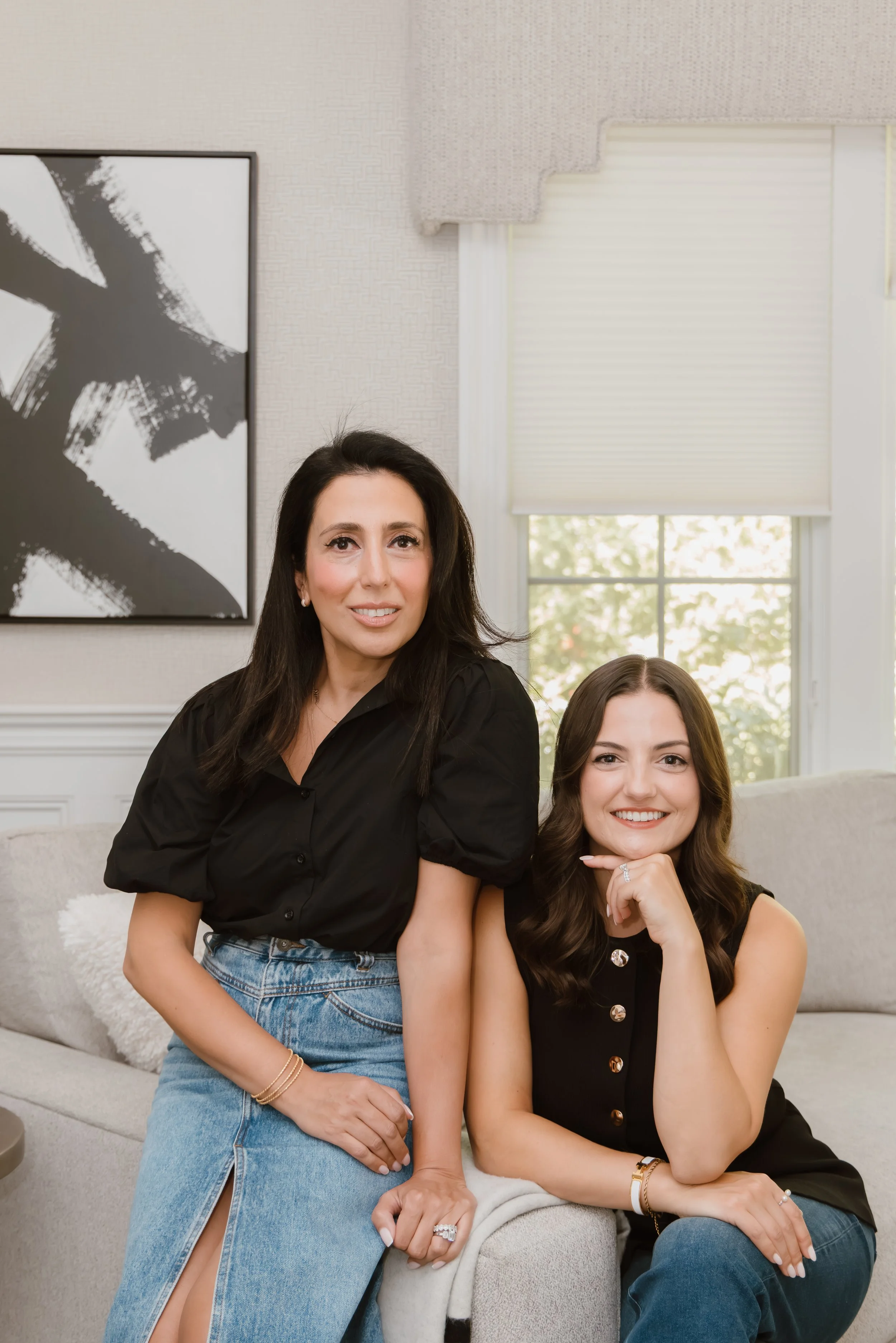 Two women sitting on a light-colored sofa in a well-lit room with abstract wall art and windows with blinds behind them.