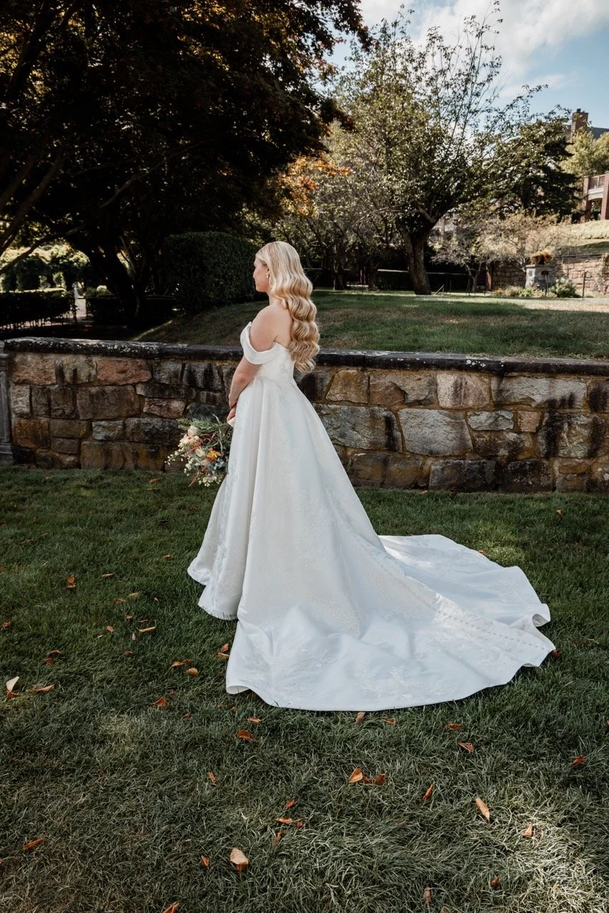 A bride in a white wedding dress holding a bouquet, standing on a grassy area outdoors with trees and a stone wall in the background.