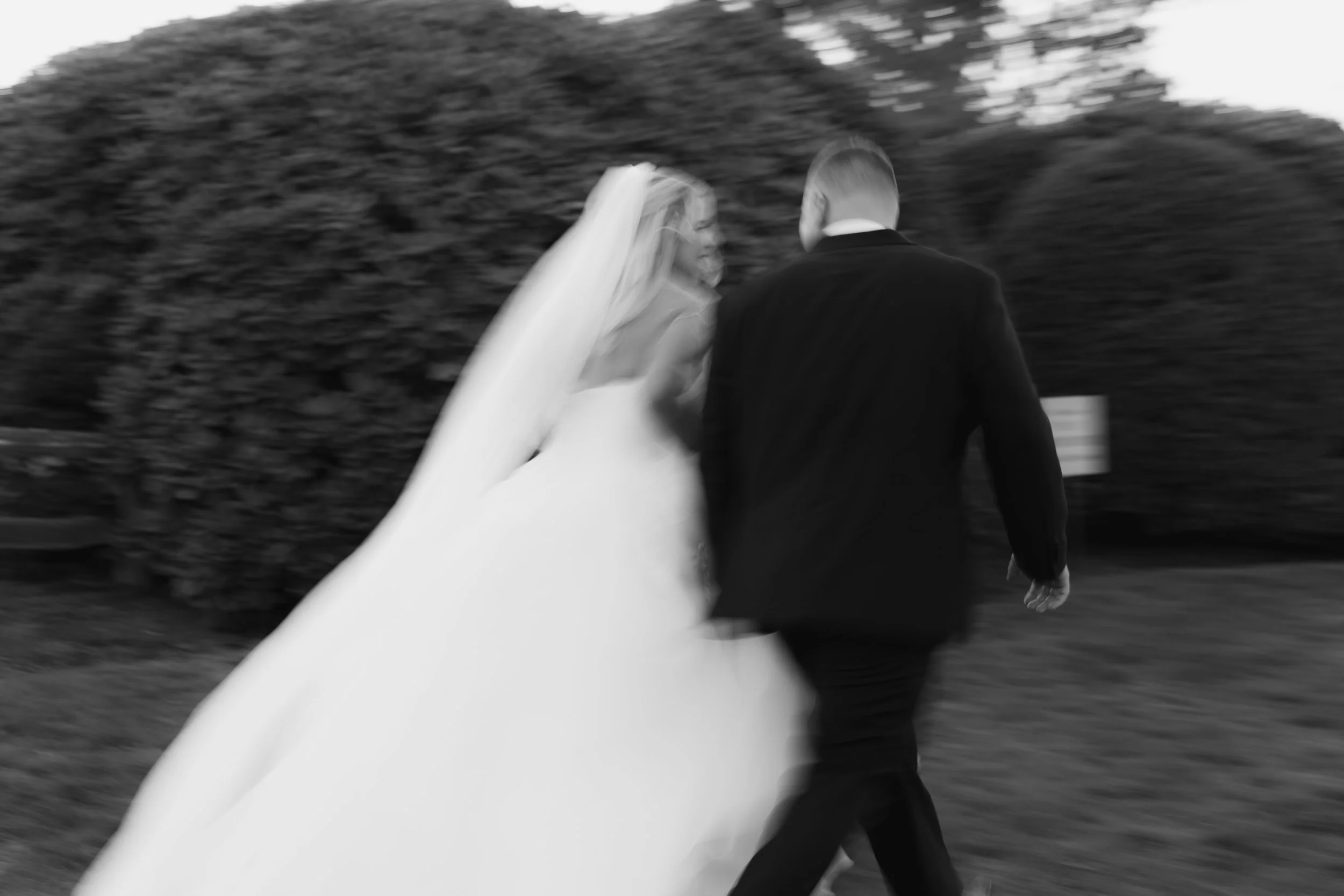A bride in a wedding dress and veil walking alongside a groom in a suit outdoors, slightly blurred, with trees in the background.